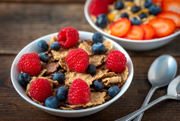 Corn flakes with blueberries and raspberries in white bowl in dark wooden desk.