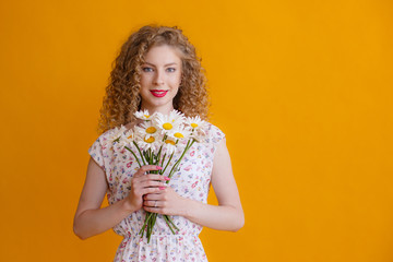 a curly blonde woman holding a bouquet of daisies on a yellow background smiles