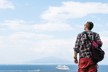 Fototapeta premium Young man with backpack standing with view on vulcano Vesuvius sea, ocean liner ship water craft. Travel and vacation concept. Copy space
