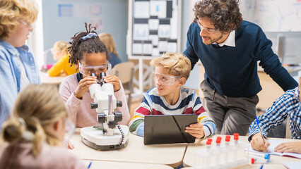 Elementary School Science Classroom: Cute Little Girl Looks Under Microscope, Boy Uses Digital Tablet Computer to Check Information on the Internet, while Enthusiastic Teacher Explains Lesson