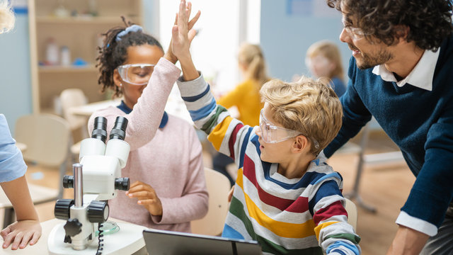 Elementary School Science Classroom: Cute Little Girl Looks Uses Microscope, Boy Uses Digital Tablet Computer to Check Information on the Internet and They do High Five in Celebration.