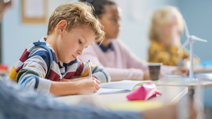 In Elementary School Classroom Brilliant Caucasian Boy Writes in Exercise Notebook, Taking Test and Writing Exam. Junior Classroom with Group of Children Working Diligently and Learning New Stuff - obrazy, fototapety, plakaty