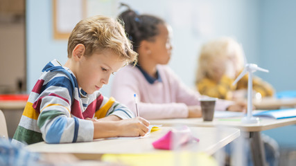 In Elementary School Classroom Brilliant Caucasian Boy Writes in Exercise Notebook, Taking Test and Writing Exam. Junior Classroom with Group of Children Working Diligently and Learning New Stuff