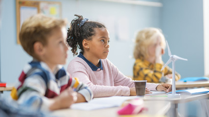 Obraz premium In Elementary School Classroom Brilliant Black Girl is Carefully Listening a Teacher. Junior Classroom with Group of Bright Children Working Diligently and Learning New Stuff