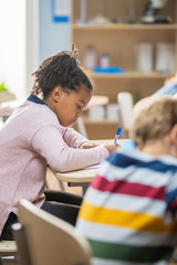 In Elementary School Classroom Brilliant Black Girl Writes in Exercise Notebook, Taking Test and Writing Exam. Junior Classroom with Group of Bright Children Working Diligently and Learning New Stuff