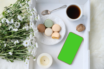 white wooden tray with black tea, coffee, plate with macaroon cakes, flowers, candles, blank screen phone, concept of technology, lifestyle, joys of life, top view