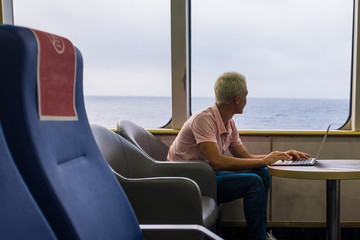 one young man using his laptop or computer pc on a ship traveling in the water of the sea or ocean - man looking outdoor while workig with device - isolated nomad alone people traveling