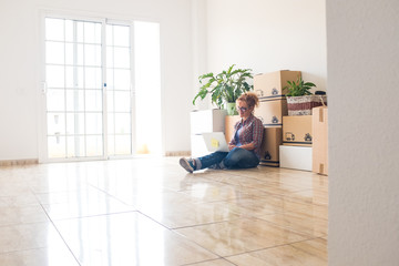 one young beautiful woman using her laptop or computer pc in a empty new house with boxes and packs on her back - independent lifestyle choosing to go live alone with a new home