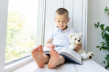 a small boy sits on the window with a book and a soft bear