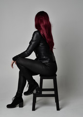 Full length portrait of  girl with red hair wearing black leather jacket, pants and boots. Sitting...