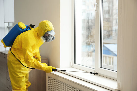 Pest Control Worker In Protective Suit Spraying Pesticide Near Window Indoors