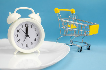 white alarm clock stands on a white plate on a blue background with a shopping cart in close-up....