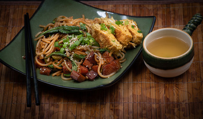 Vegan udon noodles and seitan along with  vegan omelette on square plate with chopsticks, green tea, copy space