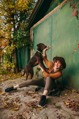 Happy girl sitting in autumn day under green wall of country house and playing with dog with smile on face. Cheerful lady in fashionable clothes playing with a dog at the cottage in the fall.