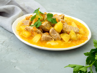 Concept of homemade food. Meat stew with potatoes and carrots. Pork meat stewed with vegetables on a grey background with grey linen towel and parsley. Tasty and nourishing lunch.
