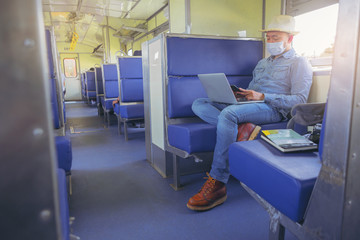 Hipster man with face mask sitting and traveling by train working on laptop in new normal style. New normal business travel concept.