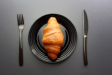 Ruddy croissant on a black plate with a fork and knife on a gray background. Crescent-shaped croissant made from puff pastry. Croissant is a popular dish in France.