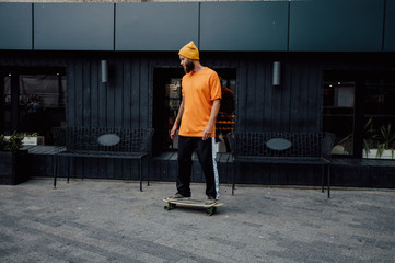 Handsome young stylish hipster guy with beard in blank orange t-shirt riding on longboard.