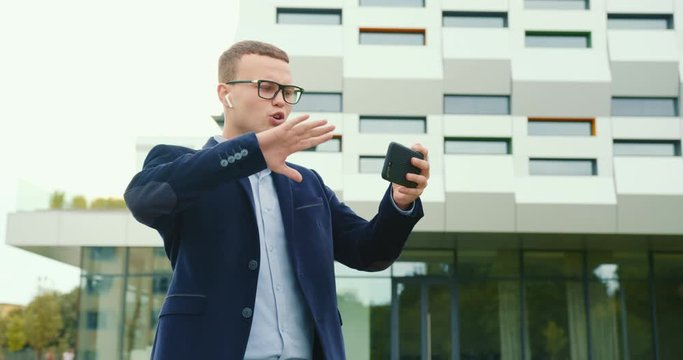 A businessman is walking near the business center and discontentedly talking via video link on the phone. He is talking in headphones. He's wearing a suit and glasses. 4K