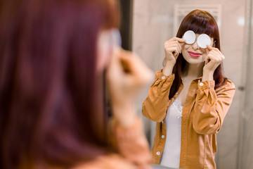 Fototapeta na wymiar Skincare routine and hygiene concept . Charming smiling Caucasian girl removing makeup, hiding her eyes with two cotton pads, standing in home bathroom and looking in mirror