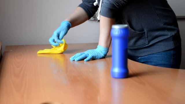 girl in a gray sweater and blue gloves washes a wooden table with a yellow rag.