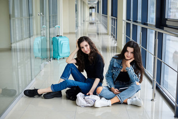 Two young brunette girls, sitting on floor in light airport hallway, with luggage behind, holding tablet, smiling, wearing casual jeans clothes. Girlfriends, traveling by air, waiting for flight.