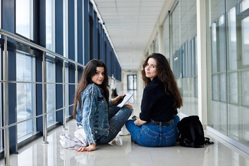 Two young brunette girls, sitting on floor in light airport hallway, with backs to camera, wearing casual jeans clothes. Girlfriends, traveling by air, waiting for flight.