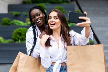 Summer sunny day. Two young women standing outdoors with shopping bags and make selfie on smartphone, looking on screen of phone.