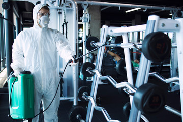 Gym disinfection and healthcare. Man in white protection suit disinfecting and fitness equipment...