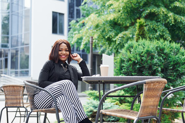 Laptop and table. Young afro american woman in fashionable clothes outdoors in the city near green trees and against business building