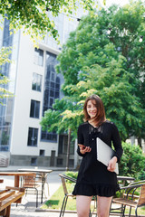 Fototapeta na wymiar Holding laptop and phone. Happy young girl in black skirt outdoors in the city near green trees and against business building