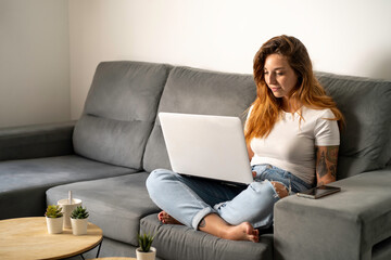 Chica joven pelirroja con pecas mirando su ordenador portátil en el sofá de su salón, relajada, tranquila y contenta, tatuada, con camiseta blanca y pantalón vaquero azul con rotos en las rodillas