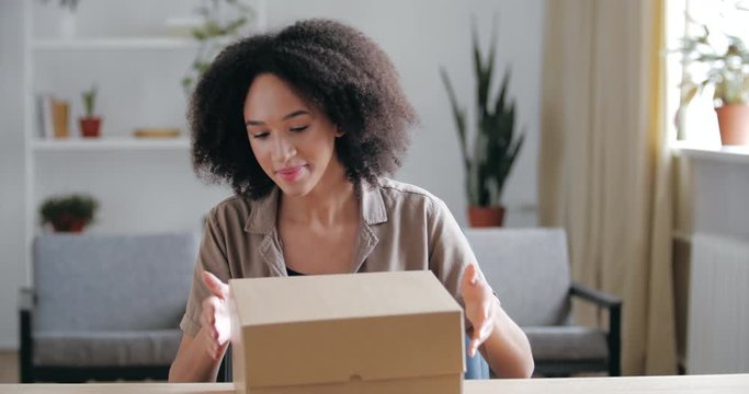 Curly brunette enters office room, brings cardboard box in her hands, puts parcel on table, unpacks Internet order gift. Young woman receives home purchase from courier, satisfied buyer customer