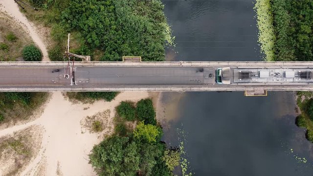 The train rides across the bridge over the river on a sunny summer day. Green trees and beach sand on the shore. Top view. Power lines