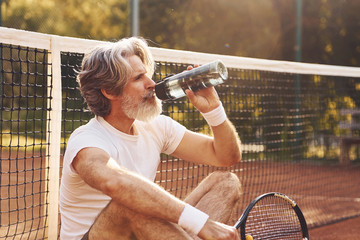 Taking break and drinking water. Senior stylish man in white shirt and black sportive shorts on tennis court