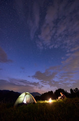 Lonely guy sitting by fire near tent on background of mountains under starry sky. Beautiful landscape of the evening sky with bright stars and clouds appearing. Night camping at summer