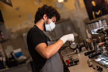 Young man, barista, making coffee espresso while wearing surgical mask and gloves for preventing corona virus spread - Bar safety working concept.