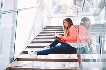 Portrait of young hipster girl reading notebook while sitting on the steps in university interior, intelligent college student in stylish clothes preparing for classes while sitting in college indoors