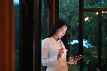 A businesswoman is holding a computer tablet and stylus pen while standing over the office glass...
