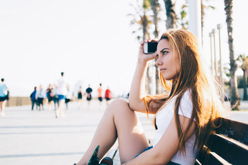 Young woman skater holding her smart phone while enjoying rest after riding on her longboard in summer day, charming female skateboarder waiting for a call on her telephone sitting on a bench