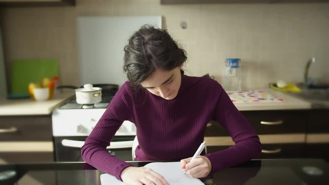 Cute student girl makes notes in a notebook, sitting at the kitchen table