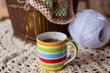 Caneca com listras coloridas, cheia de chá, sobre a mesa com novelos e crochê ao fundo.