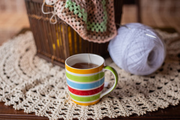 Caneca com listras coloridas, com chá, sobre a mesa com novelos e crochê ao fundo.