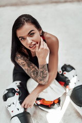 Fototapeta na wymiar Beautiful looking woman with tattoos in a black and red tracksuit, white boots and with a motorcycle helmet, posing in a white photo studio, sitting on the floor