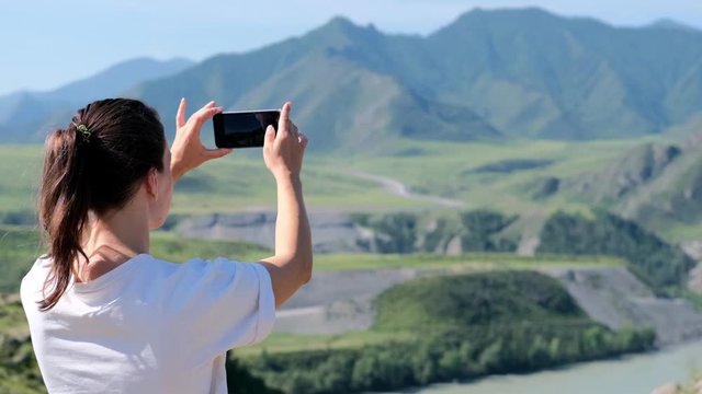 Back view of a girl photographing a beautiful landscape on a smartphone. Cinematic and inspiring travel blogger lives a motivational adventure. Happy young woman in mountains
