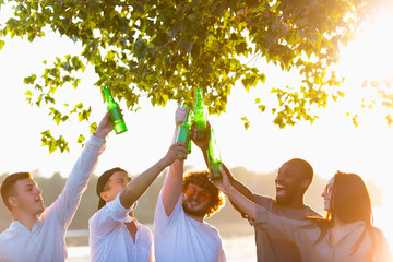 Warm. Group of friends clinking beer bottles during picnic at the beach in sunshine. Lifestyle,...