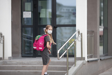 A schoolgirl wearing medical face mask and carrying a pink backpack goes up the stairs of her school, looking back to her parents. Back to school. Protection from virus for school.