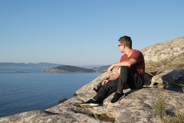 Naklejka na ściany i meble Boy sitting on the rocks of a mountain looking at the ocean