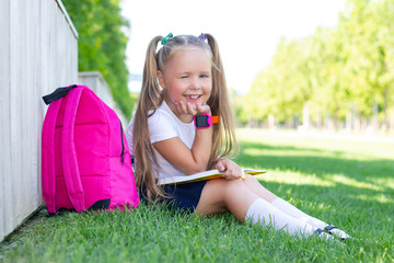 schoolgirl sits on the grass with a backpack and a textbook and smiles.