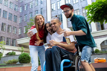 Fototapeta na wymiar Selfie time. Group of friends taking a stroll on city's street in summer day. Handicapped man with his friends having fun. Inclusion and diversity concept, normal lifestyle of special groups of